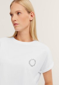 Weißes Baumwoll-T-Shirt mit aufgerollten Ärmeln und einem kreisförmigen schwarzen Druck mit Text auf der linken Brustseite. Glatte Textur, klassischer Schnitt.
