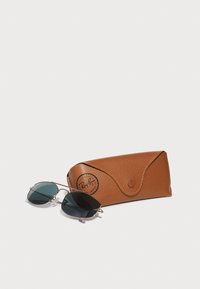 Ray-Ban'i prillid tumedate läätsedega ja õhukese metallraamiga, mis on asetatud tekstuurse pruuni nahksehitsa kõrvale. Sees on suurepäraselt reljeefne logo ja nööpkinnitus.