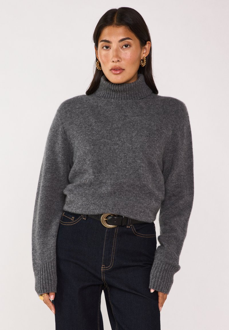 Maglione grigio a collo alto con polsini e orlo a coste, realizzato in materiale morbido e testurizzato, abbinato a jeans scuri e una cintura decorativa.