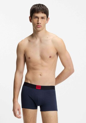 Młody mężczyzna z krótkimi ciemnymi włosami, ubrany w granatowe bokserki z czarną gumką i czerwoną metką "Hugo", stojący na jednolitym białym tle.