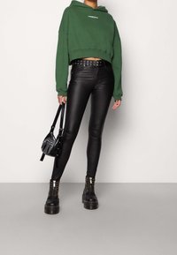 Persona che indossa un crop hoodie verde, pantaloni in pelle neri a vita alta, cintura borchiata, stivali neri chunky con lacci, e tiene in mano una borsa nera.