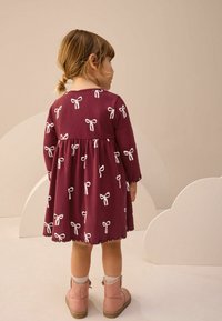 Mažylė mergaitė su rudais plaukais, surištais į uodegėlę, vilkinti bordo suknelę su baltais kaspinais ir rožiniais batais, stovi priešais smėlio spalvos foną.