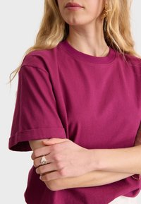 Majica kratkih rukava s okruglim izrezom u jednoličnoj magenta boji, izrađena od mekane pamučne tkanine s povijenim rukavima i opuštenim krojem. Jednostavan dizajn.