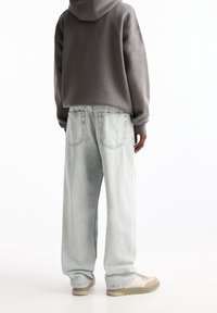 Person wearing a gray hoodie, light washed wide-leg jeans, and beige sneakers, standing facing away against a white background.