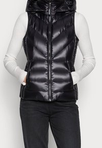 Zwarte pufferbodywarmer met capuchon, voorzien van een glanzende afwerking, visgraatstiksels, een ritssluiting aan de voorkant en zijzakken. Draagt over een wit shirt met lange mouwen.