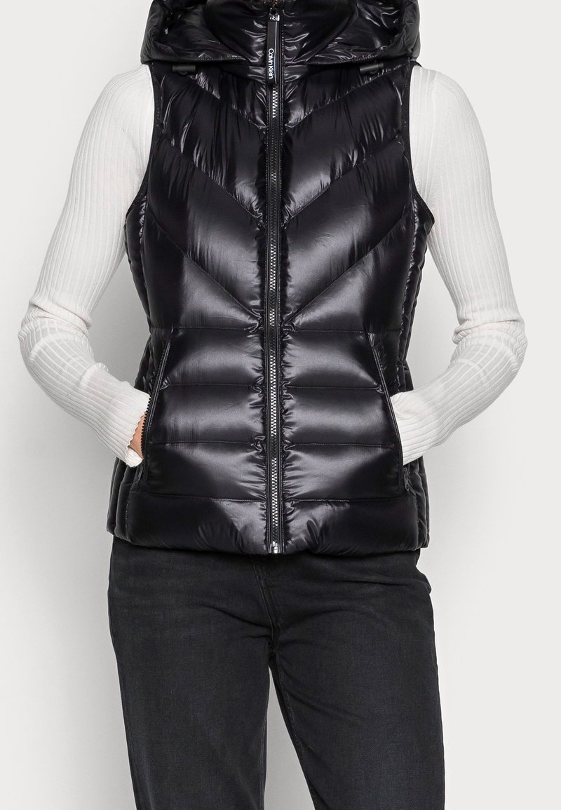 Zwarte pufferbodywarmer met capuchon, voorzien van een glanzende afwerking, visgraatstiksels, een ritssluiting aan de voorkant en zijzakken. Draagt over een wit shirt met lange mouwen.