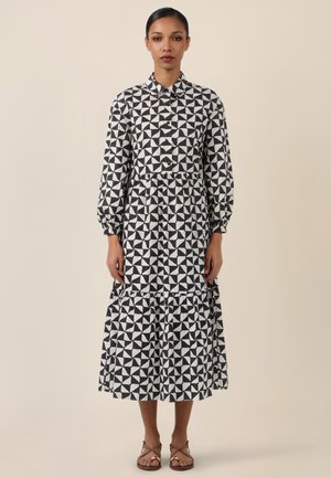 Woman stands wearing a long-sleeve, black and white geometric patterned midi dress with buttoned front and tiered skirt, paired with flat sandals.