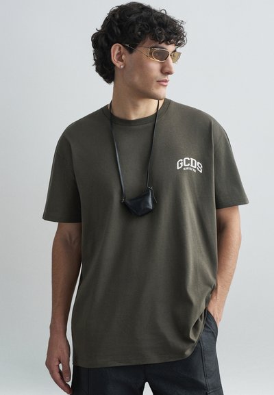 T-shirt en coton vert olive à manches courtes, avec un logo "GCDS" blanc sur la poitrine, porté avec un petit collier en pochette noire.
