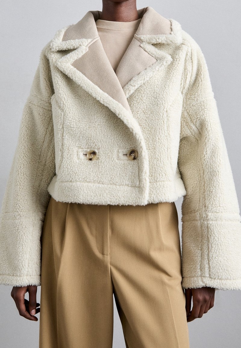 Vue rapprochée d'une personne portant une veste sherpa crème raccourcie avec de grands revers et des boutons, associée à un pantalon taille haute beige.