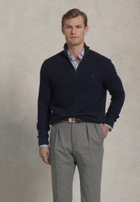 Ung mann iført en marineblå quarter-zip genser over en stripete skjorte og grå plissert bukser, som står mot en enkel lys bakgrunn.