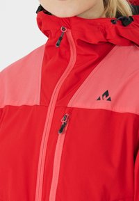 Veste imperméable rouge avec un col montant, panneaux contrastants roses, fermeture éclair et détail de logo noir sur la poitrine. Texture lisse.