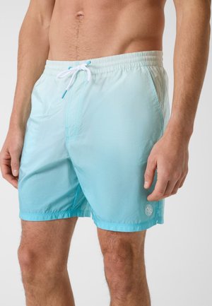 Mann trägt hellblaue Badeshorts mit weißem Kordelzug, steht mit entspannten Armen an den Seiten vor einfarbigem Hintergrund.