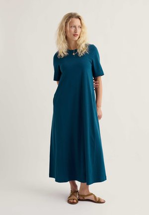 Szőke nő türkiz rövid ujjú maxiruha és barna szandálok viselésével, egyik kezét a háta mögött tartva áll.