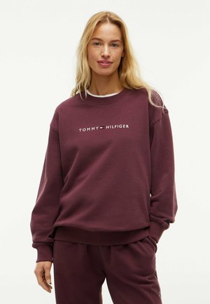 Lyshåret kvinde iført en vinrød Tommy Hilfiger sweatshirt og matchende bukser, stående med den ene hånd i lommen mod en ensfarvet baggrund.