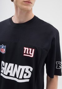 T-shirt nero della NFL con la scritta "GIANTS" in grande carattere bianco, logo "ny" in rosso e scudetto NFL sul lato sinistro del petto; maniche corte e collo a giro.
