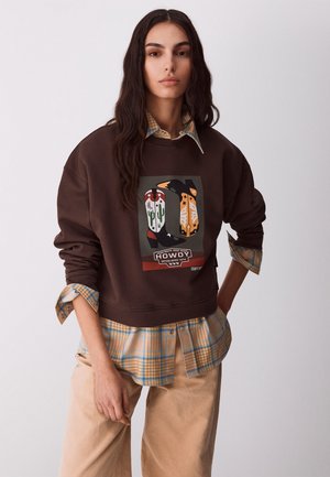 Młoda kobieta z długimi ciemnymi włosami, ubrana w brązową bluzę z grafiką kowbojskich butów i warstwową koszulę w kratę, beżowe spodnie, pozująca na białym tle.