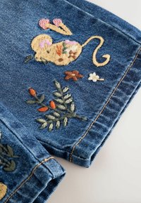 Denimstoff mit buntem gesticktem Mausmotiv und verschiedenen Blumen und Blättern in pink, orange, braun, grün und creme farbigen Fäden.