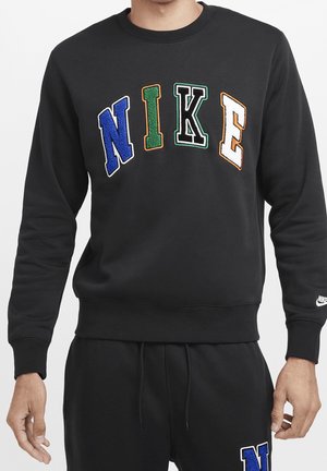 Mand iført sort sweatshirt og bukser med store flerfarvede "NIKE" bogstaver på sweatshirt og lille hvid Nike-logo på ærmet.