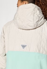 Podložena jakna v bež barvi z nižjim delom v metini zeleni, z kapuco in logotipom na hrbtu, ki prikazuje prešito teksturo.