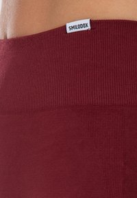 Bordeauxrode geribbelde tailleband met een gladde textuur. Klein 'SMILODOX'-label aan de bovenkant. De stof lijkt rekbaar en aansluitend te zijn.