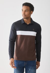 Man wearing long-sleeve polo shirt with black, white, and brown color blocks, paired with blue jeans, looking slightly to the side.