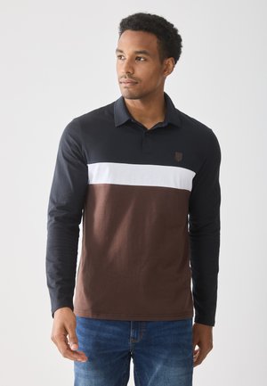 Man in een poloshirt met lange mouwen in zwart, wit en bruin kleurblokken, gecombineerd met blauwe jeans, kijkt iets opzij.