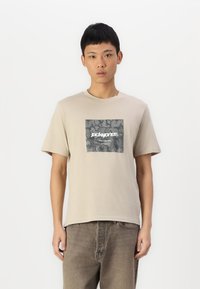 Camiseta de algodón beige de manga corta, con un estampado gráfico cuadrado gris y texto blanco, combinada con pantalones vaqueros de color marrón claro.