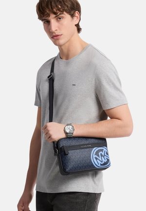 Mladić u svijetlosivoj majici i srebrnom satu, drži tamnoplavu Michael Kors torbu s crnim remenom.