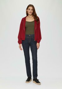 Czerwona bluza z zamkiem na całej długości, oliwkowa koszulka na ramiączkach, ciemnoniebieskie dżinsy i czarne sneakersy. Gładki materiał, luźny krój, widoczne detale szycia.