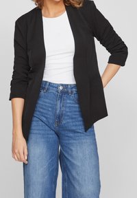 Svart kavaj med öppen framsida, strukturerade axlar och trekvartsärmar, i kombination med en vit topp och högmidjade blå jeans.