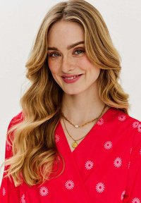 Červené šaty s bílými květinovými vzory, s výstřihem do V a vrstevnými zlatými náhrdelníky s perlovými a medailonovými akcenty.