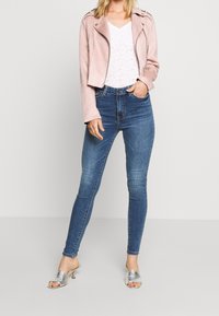 Růžová semišová moto bunda s krátkým střihem přes bílý krajkový top, doplněná vysokými modrými skinny džíny a stříbrnými sandály na podpatku.