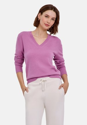 Femme aux cheveux courts et bruns portant des boucles d'oreilles créoles dorées, un pull violet à col en V et un pantalon blanc à cordon, les mains dans les poches.