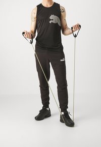 Débardeur noir avec un motif gris, pantalon de survêtement noir avec logo, et chaussures de sport noires. Tenant des bandes de résistance avec poignées.