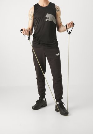 Černý tílko se šedým motivem, černé tepláky s logem a černé sportovní boty. Drží odporové pásy s úchyty.