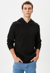 Zwarte hoodie met een trekkoordkap, lange mouwen en geribbelde manchetten en zoom. Gemaakt van zachte stof, gedragen met lichtblauwe jeans.