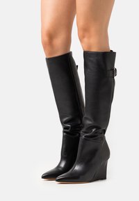 Botas de cuña negras hasta la rodilla con puntera afilada y correa de hebilla lateral, usadas sobre piernas desnudas contra un fondo blanco.