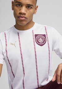 Camisa desportiva branca com riscas verticais em carmesim, logo dourado da Puma e o emblema do Manchester City no lado esquerdo do peito. Mangas curtas e gola redonda.
