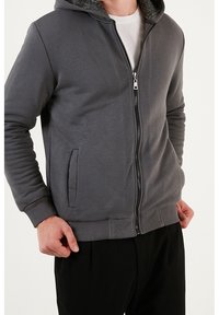 Felpa grigia con zip, dotata di cappuccio foderato, tasche laterali e polsini a coste. Realizzata in morbido tessuto testurizzato, presenta una zip argentata.