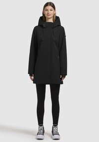 Veste imperméable noire avec un col haut et une capuche ajustable, dotée de deux poches avant et d'une coupe droite. Portée sur des leggings noirs et des baskets.