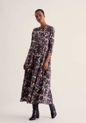 Femme portant une longue robe fleurie à manches trois-quarts et des bottes noires à talons, debout devant un fond clair uni.