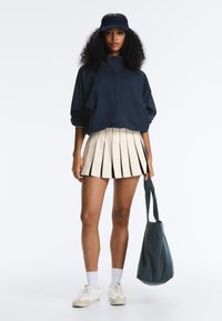 Nő tengerészkék napellenzőt és dzsekit visel, krémszínű pliszírozott szoknyát, fehér zoknit és sportcipőt, sötét színű nagy táskát tart az oldalán.