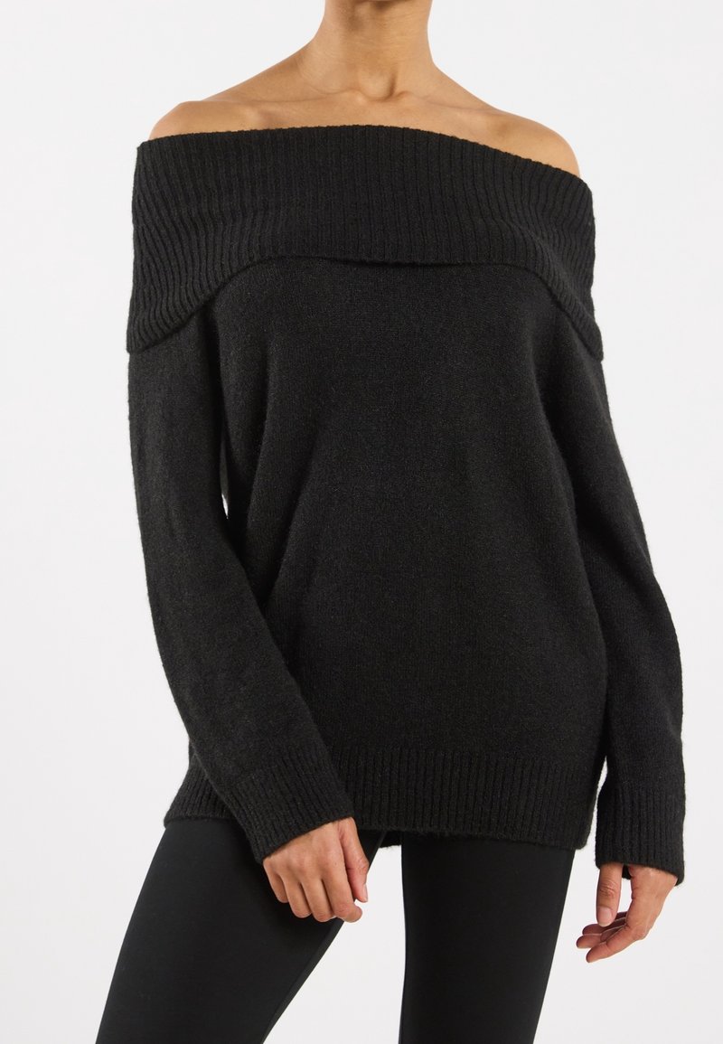 Femme portant un pull côtelé noir à épaules dénudées avec manches longues et un legging noir, debout devant un fond blanc uni.