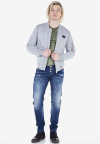 Grijze bomberjack met ribgebreide manchet, rits aan de voorkant en logo-opdruk, gelaagd over een groene T-shirt, gecombineerd met versleten blauwe jeans en zwarte laarzen.