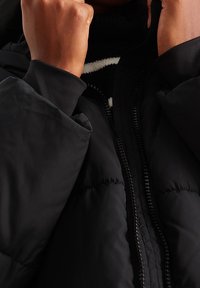 Czarna kurtka ocieplana z zamkiem błyskawicznym, z wysokim kołnierzem. Materiał wydaje się gładki o miękkiej teksturze i minimalnych detalach szwów.