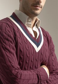 Jersey de punto cable en color burdeos con un profundo escote en forma de V; presenta acentos en blanco y azul marino alrededor del cuello. Lleva una camisa con cuello debajo.