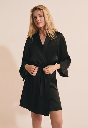 Blonde Frau, die einen schwarzen Satinkleid mit weiten Ärmeln und gebundenem Taillengürtel richtet, vor einem beigen Hintergrund stehend.