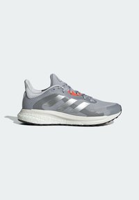 Chaussure de running Adidas grise avec des bandes argentées, une semelle blanche rembourrée et des œillets de lacet orange sur un fond clair.