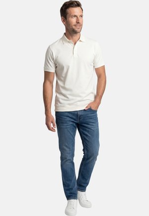 Man die een witte polo met korte mouwen, blauwe spijkerbroek en witte sneakers draagt, staat met één hand in de zak en kijkt opzij.