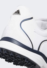 Chaussure de sport blanche avec une tige texturée, des accents argentés et une semelle bleue marine. Présente des motifs complexes et une semelle extérieure en caoutchouc moulé pour une bonne adhérence.